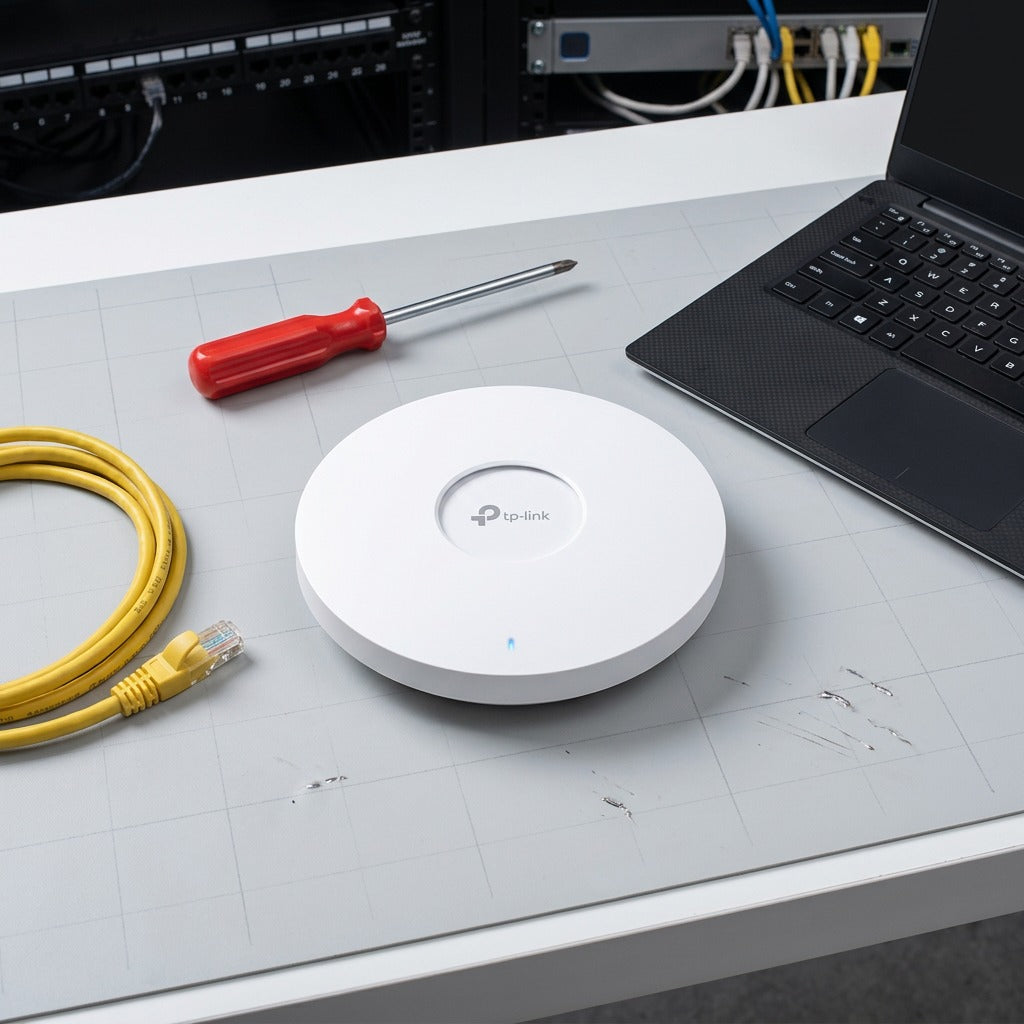 TP-Link EAP670 Wi-Fi 6 access point shown on a workbench next to a laptop, screwdriver, and cable.