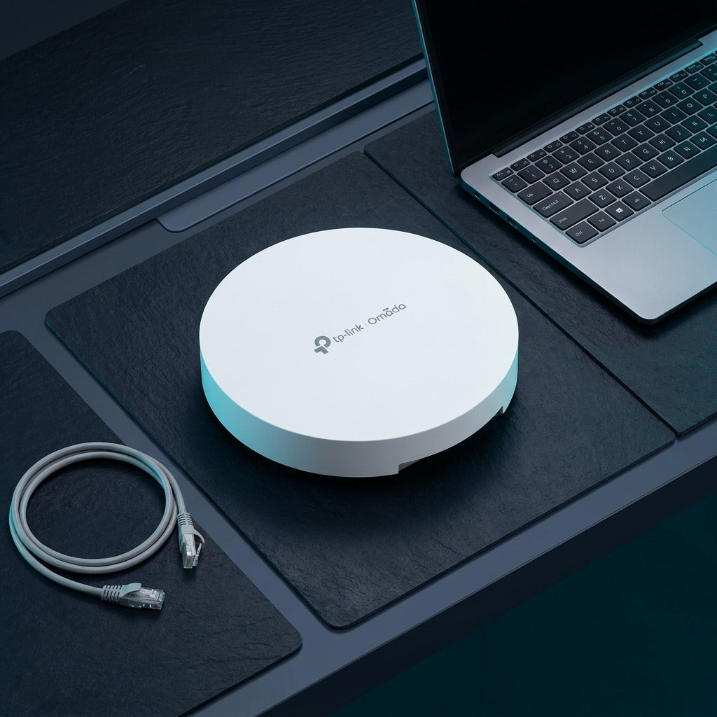 White TP-Link Omada Wi-Fi 7 access point on a dark workbench next to a laptop and ethernet cable.